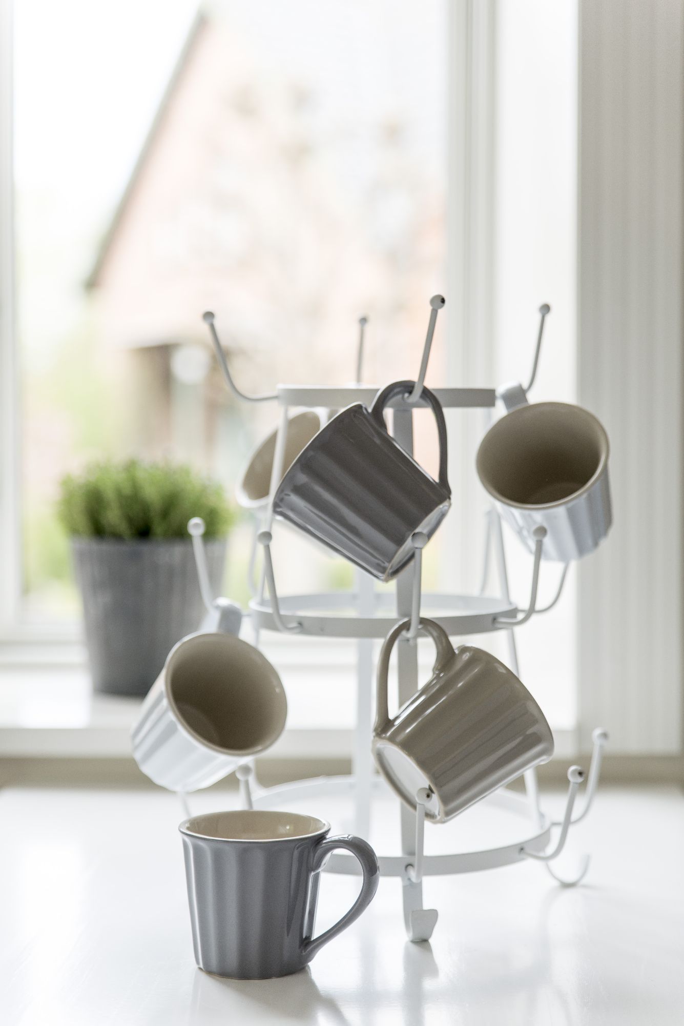 Ein Kaffeetassenständer mit mehreren Tassen in verschiedenen Grautönen, im Hintergrund ein Fenster mit Pflanzen.
