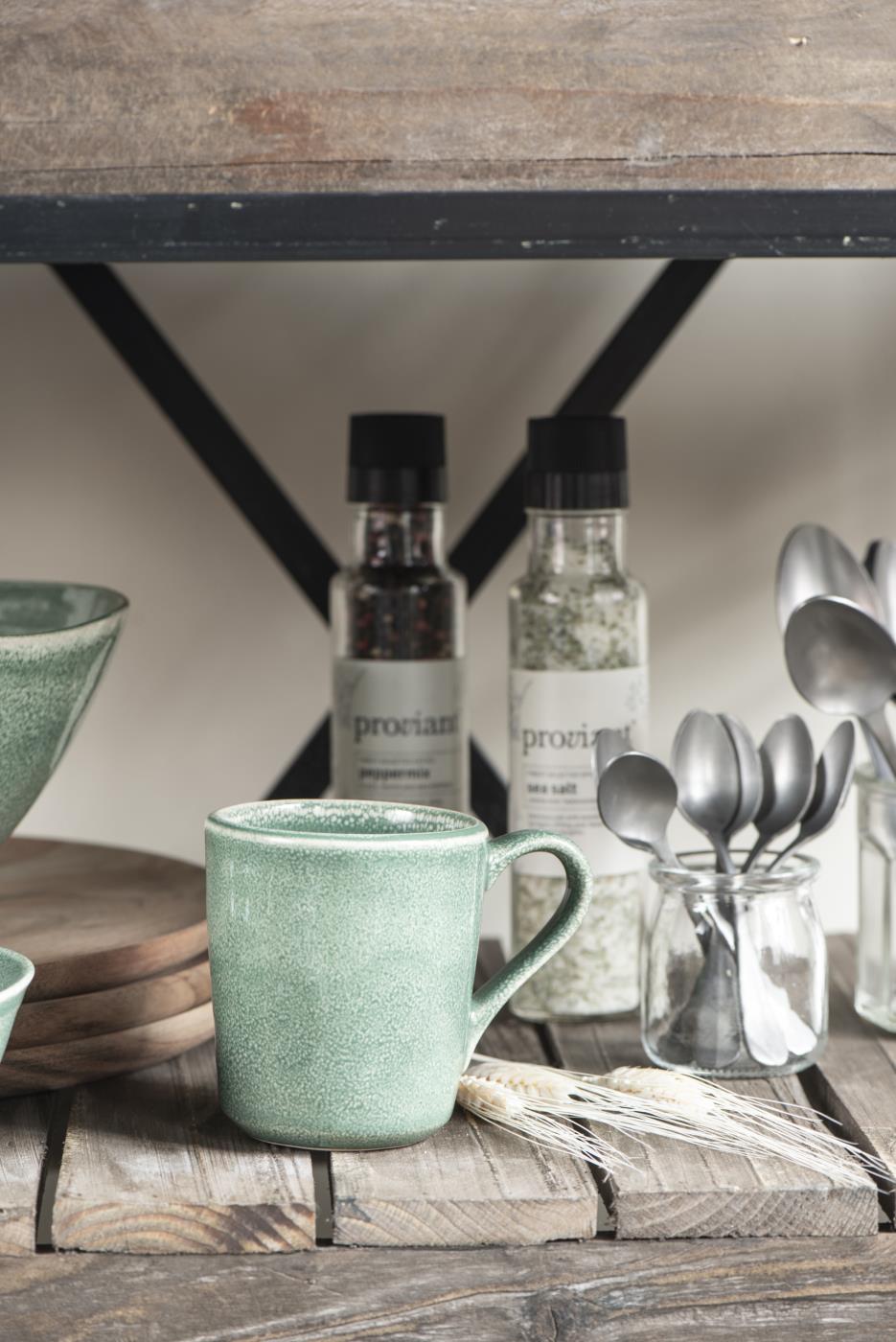 Eine grüne Tasse steht auf einem Holzregal. Im Hintergrund sind Gewürzgläser und Löffel in einem Glas zu sehen.