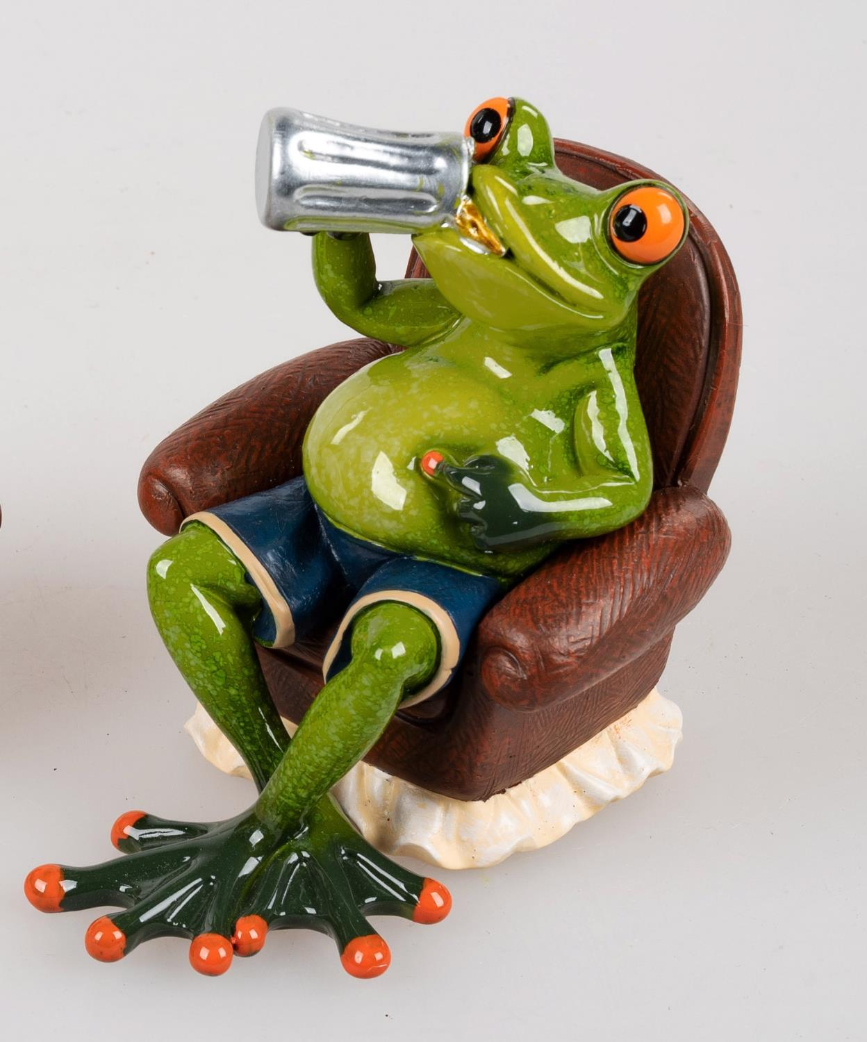 Eine grüne Froschfigur sitzt in einem Sessel, trägt Shorts und hält ein Glas in der Hand. Die Augen