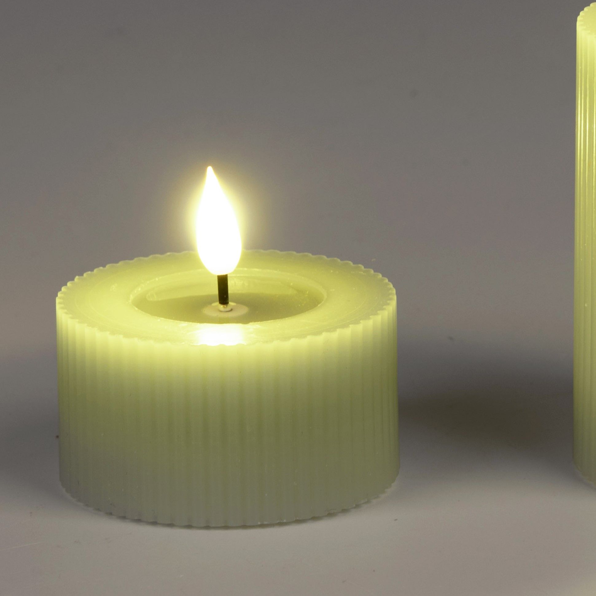 Zwei grüne LED-Kerzen, eine mit flackernder Flamme, stehen nebeneinander auf einem hellen Untergrund
