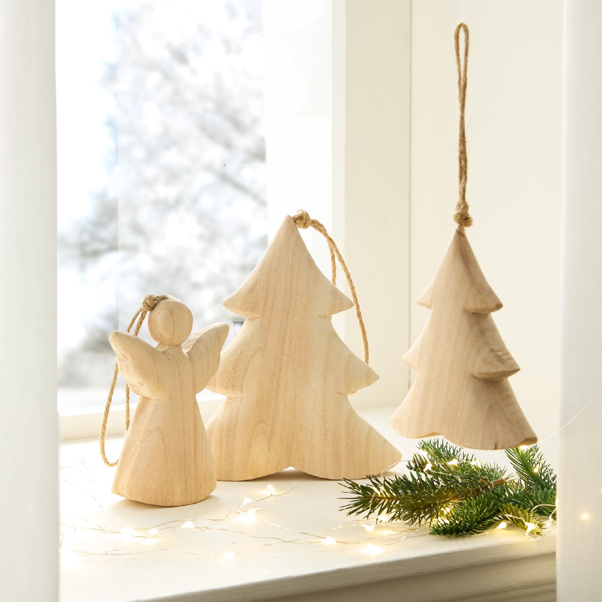 Drei Holzfiguren: ein Engel und zwei Weihnachtsbäume, stehen auf einer Fensterbank, umgeben von Lich