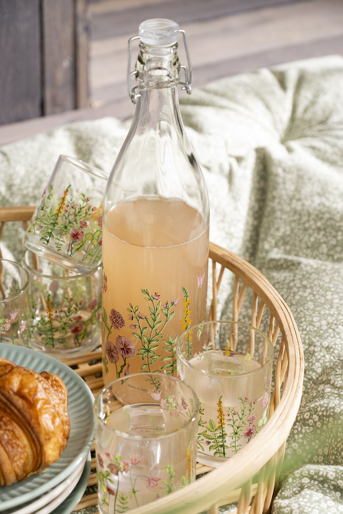 Ein Korb mit einer Glasflasche und mehreren Gläsern, die mit Blumenmotiven verziert sind. Ein Croissant und Teller sind ebenfalls sichtbar.