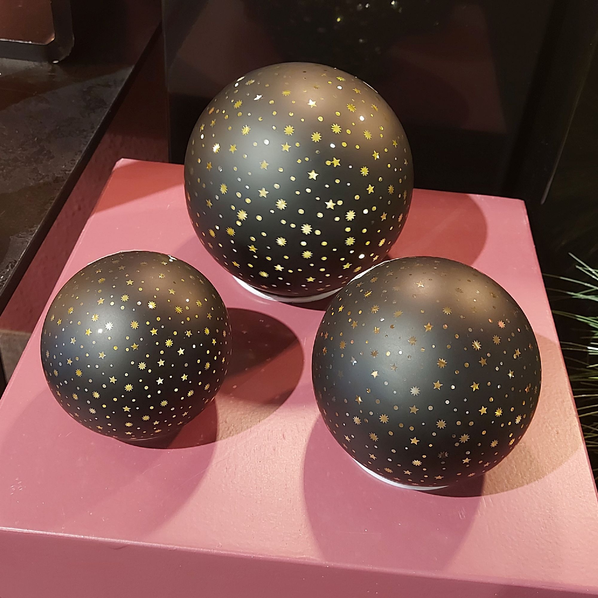 Drei schwarze Kugeln mit goldenen Sternenmustern, unterschiedlich groß, auf einer rosa Plattform ang