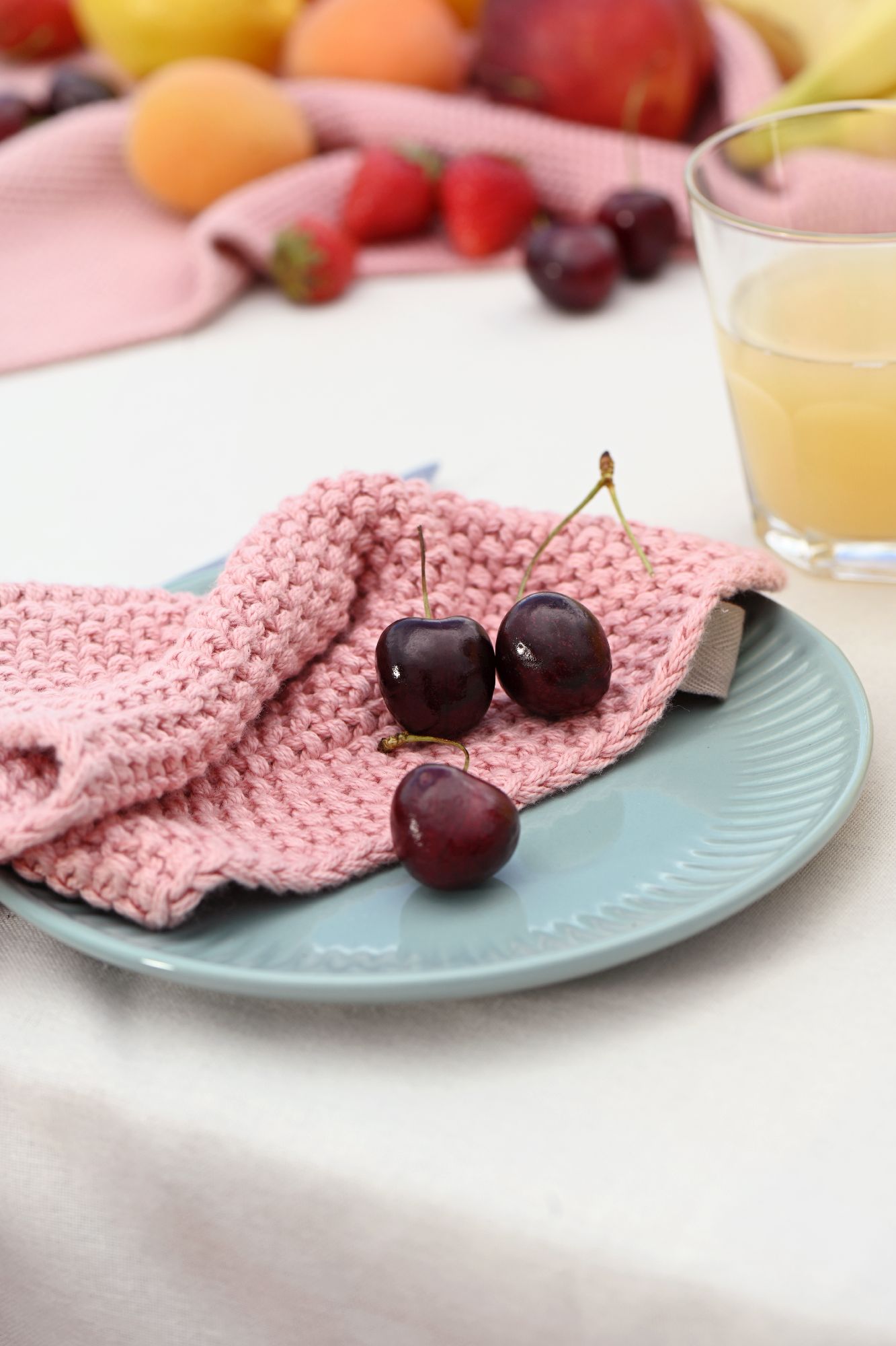 Ein blauer Teller mit drei Kirschen und einem rosa Geschirrtuch. Im Hintergrund sind verschiedene Früchte und ein Glas Saft sichtbar.