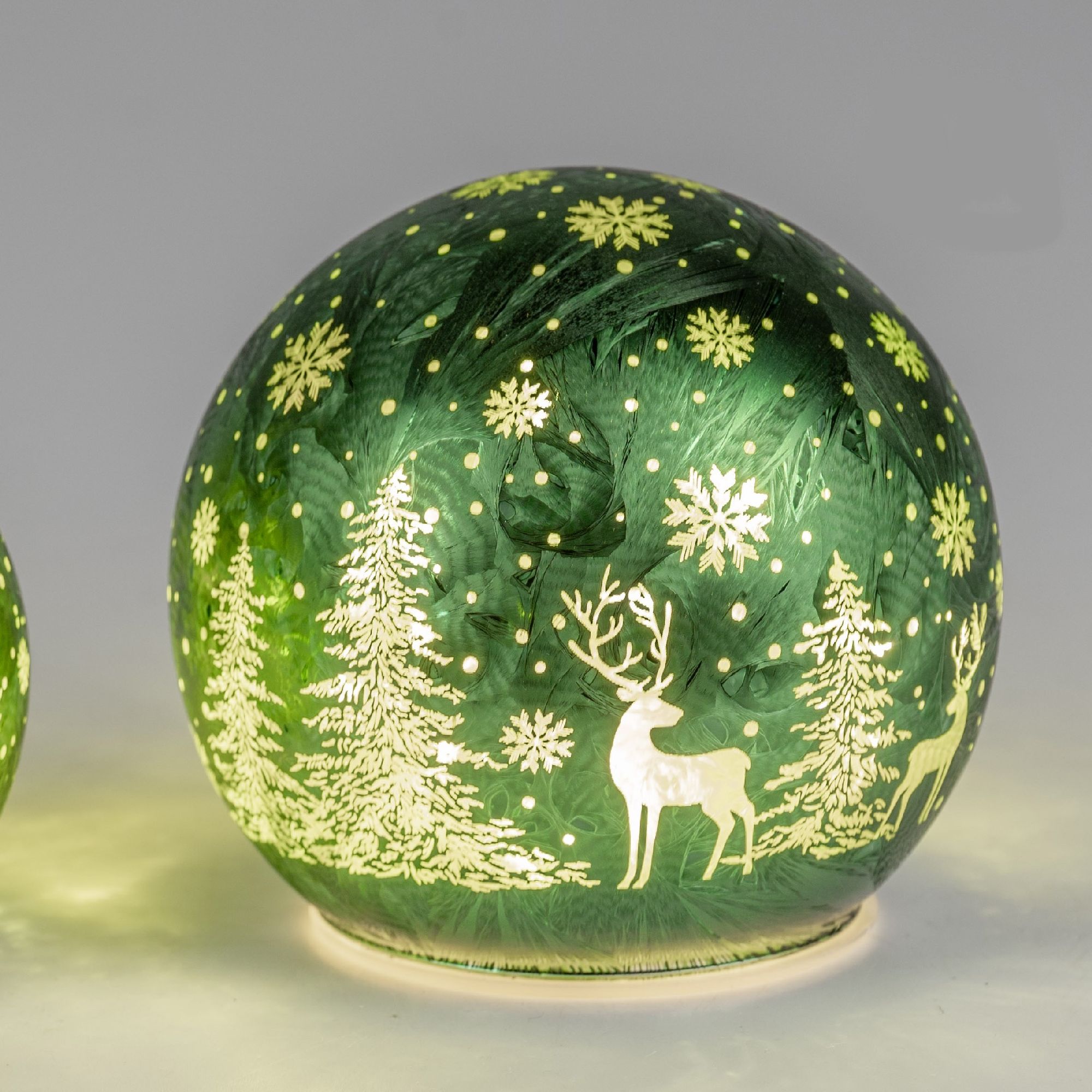 Grüne Glaskugel mit weißen Motiven von Bäumen, Schneeflocken und Rentieren, auf hellem Hintergrund.
