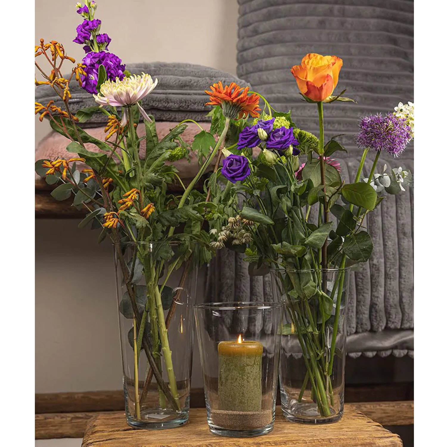 Drei Glasvasen mit bunten Blumen und eine Kerze auf einem Holztablett, im Hintergrund ein graues, ge