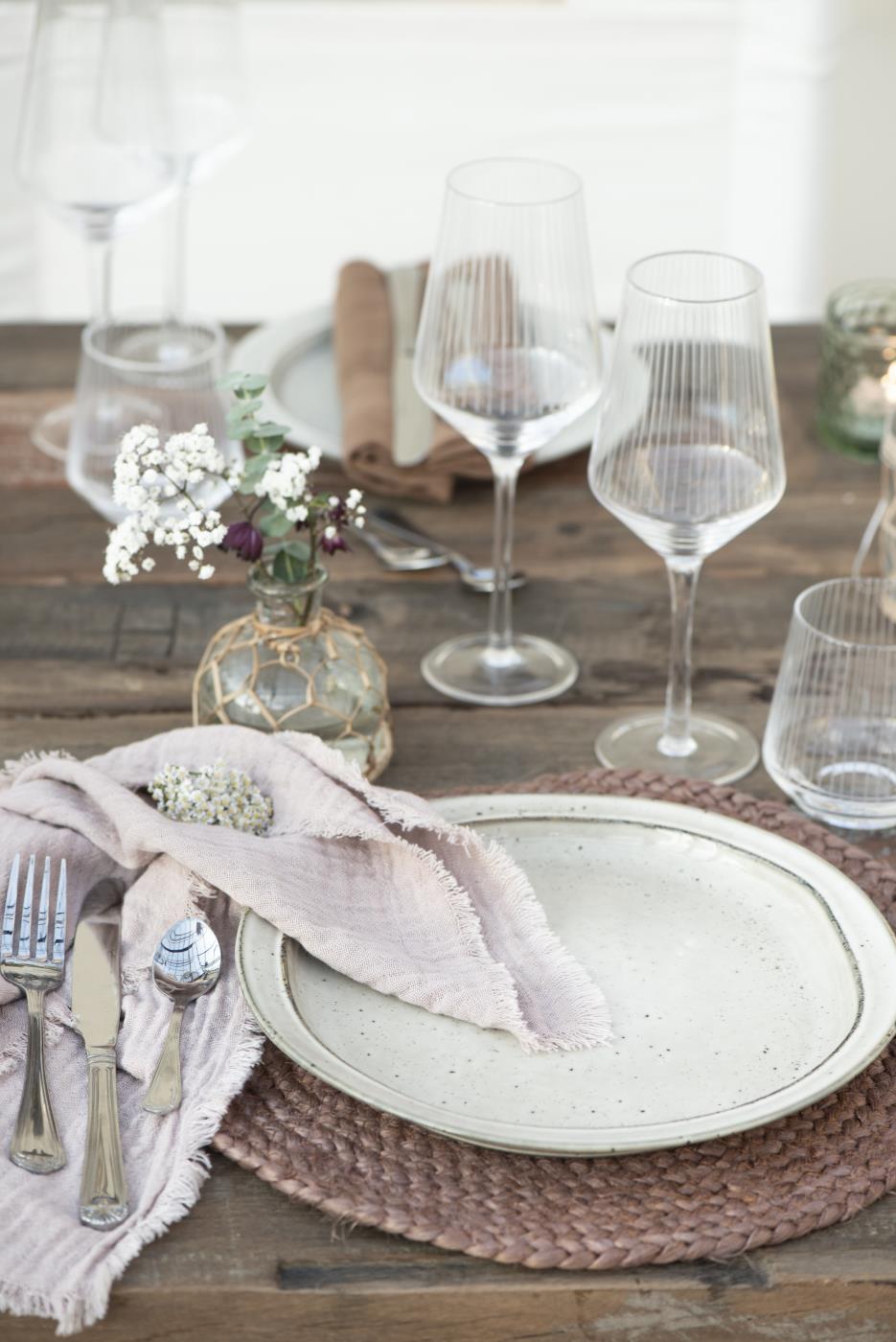 Ein gedeckter Tisch mit einem Teller, Besteck, Gläsern und einer kleinen Blumendekoration in einer Glasvase.