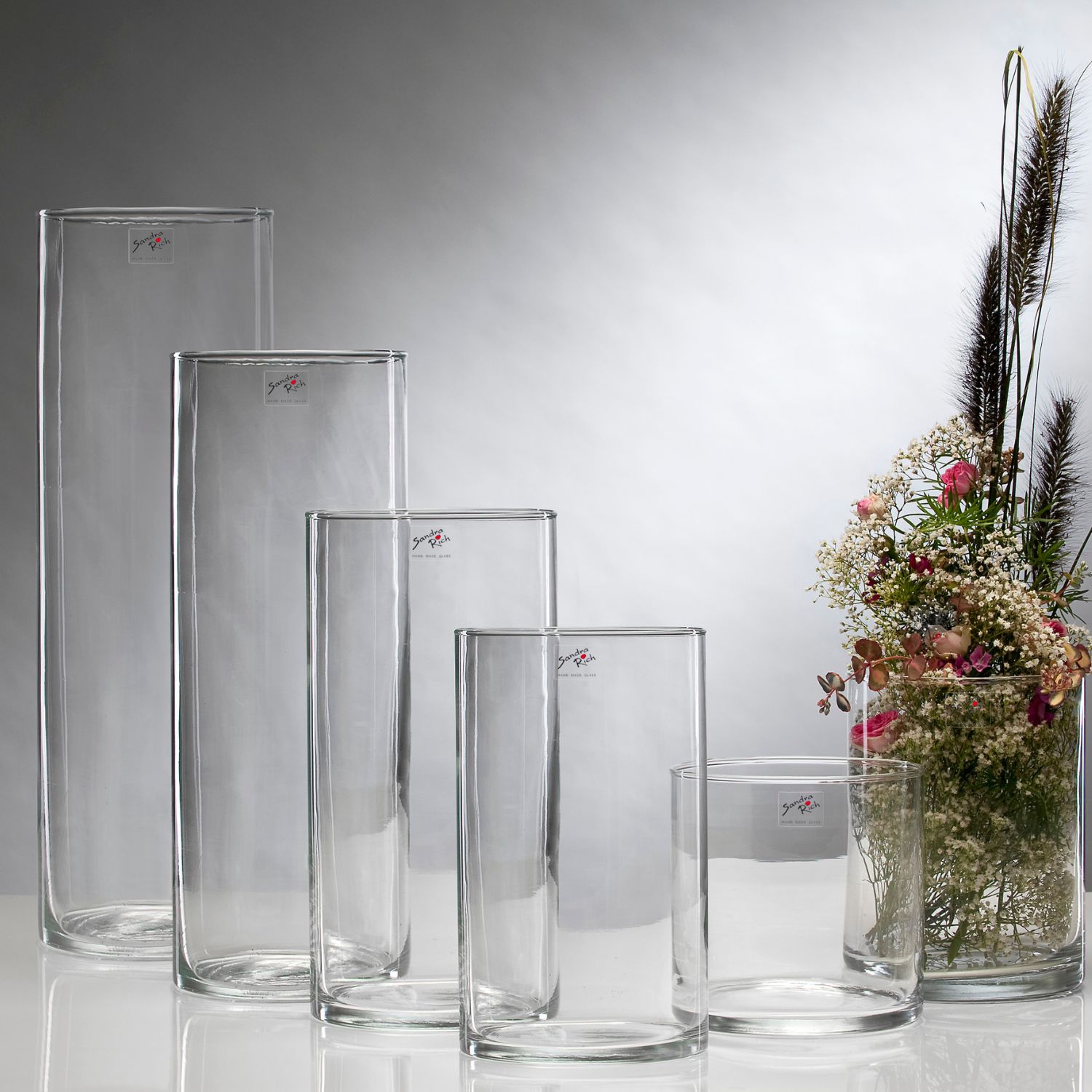 Fünf transparente Glasvasen in unterschiedlichen Höhen, eine Vase mit bunten Blumen und Gräsern. Hintergrund in sanften Grautönen.