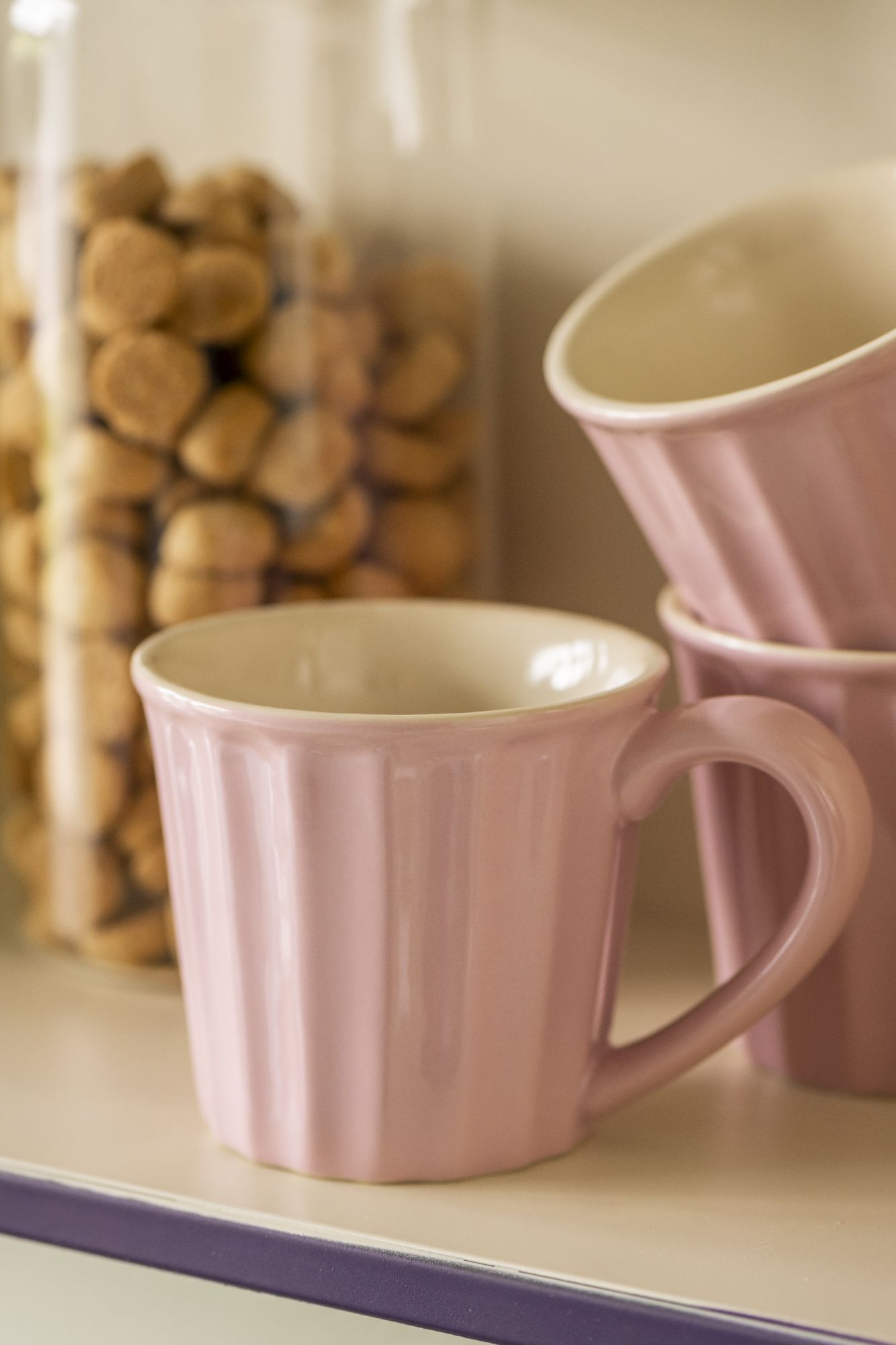 Eine rosa Tasse steht auf einem Regal, daneben sind weitere Tassen und ein Glas mit runden Keksen zu sehen.