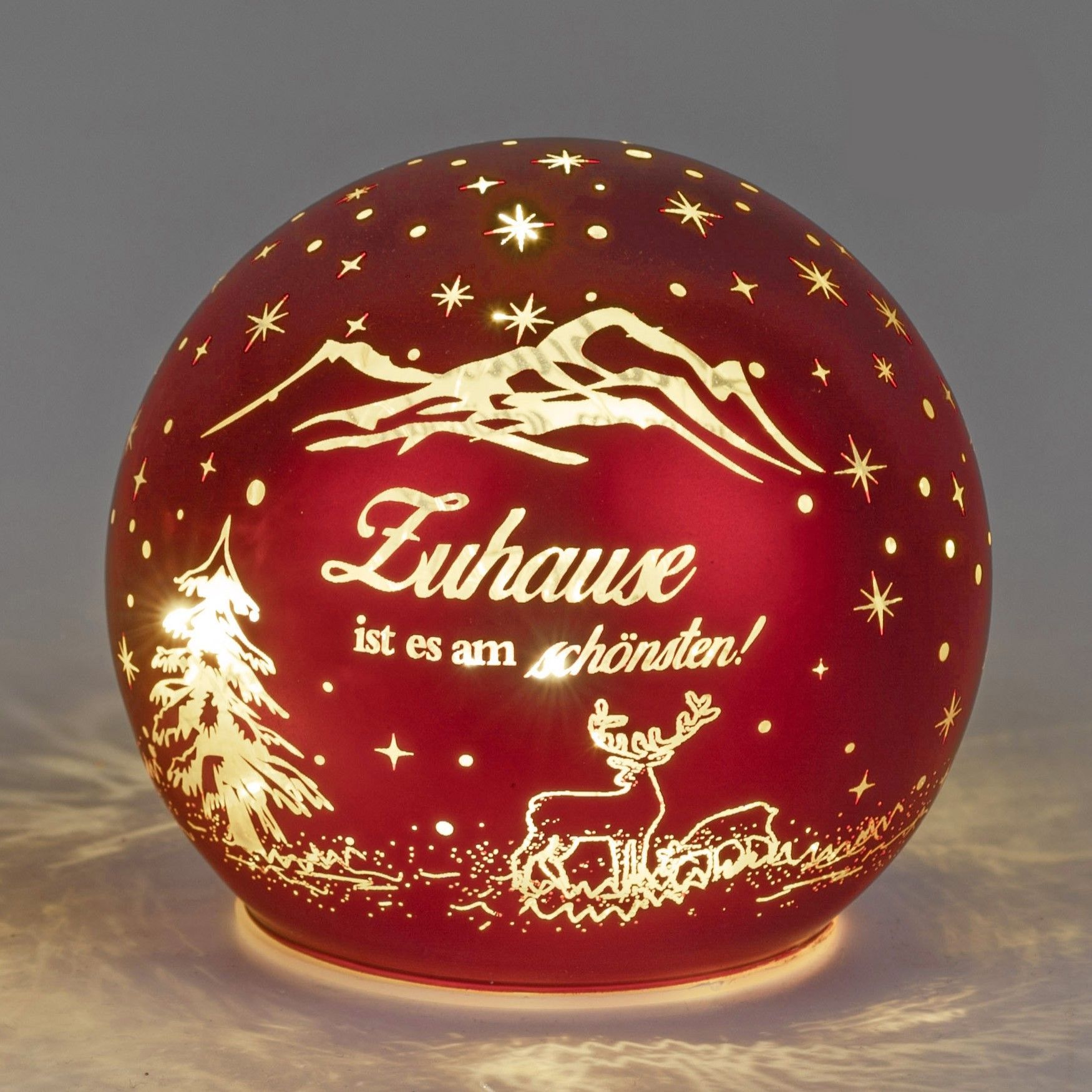 Rote Glaskugel mit Weihnachtsmotiven, darunter ein Baum, Berge und der Schriftzug „Zuhause ist es am