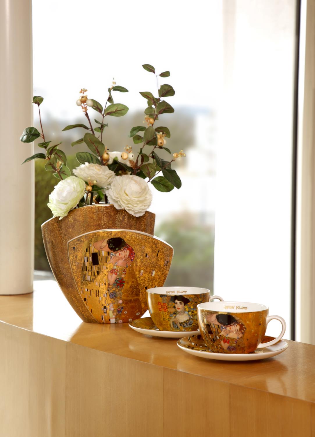 Vase mit Blumen und zwei Tassen auf einem Tisch. Die Tassen sind mit goldenen Mustern verziert. Heller Hintergrund.