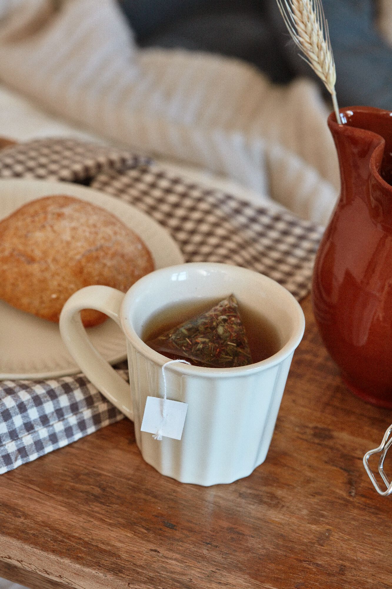 Eine Tasse mit Kräutertee und einem Teebeutel steht auf einem Holztablett. Daneben liegt ein Brötchen und ein kleiner Krug.