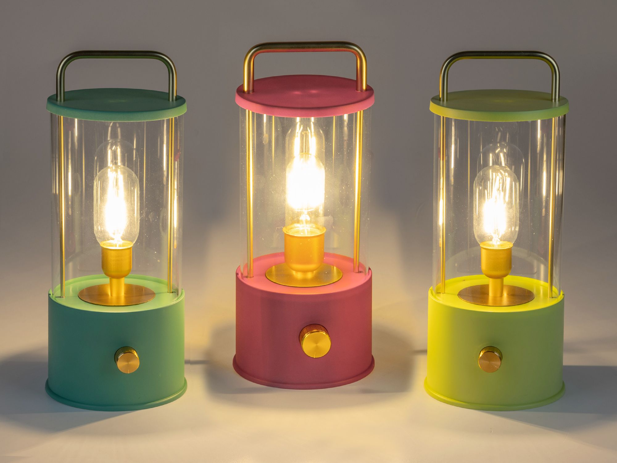 Drei Tischlampen in verschiedenen Farben (grün, pink, gelb) mit transparenten Glaskörpern und golden