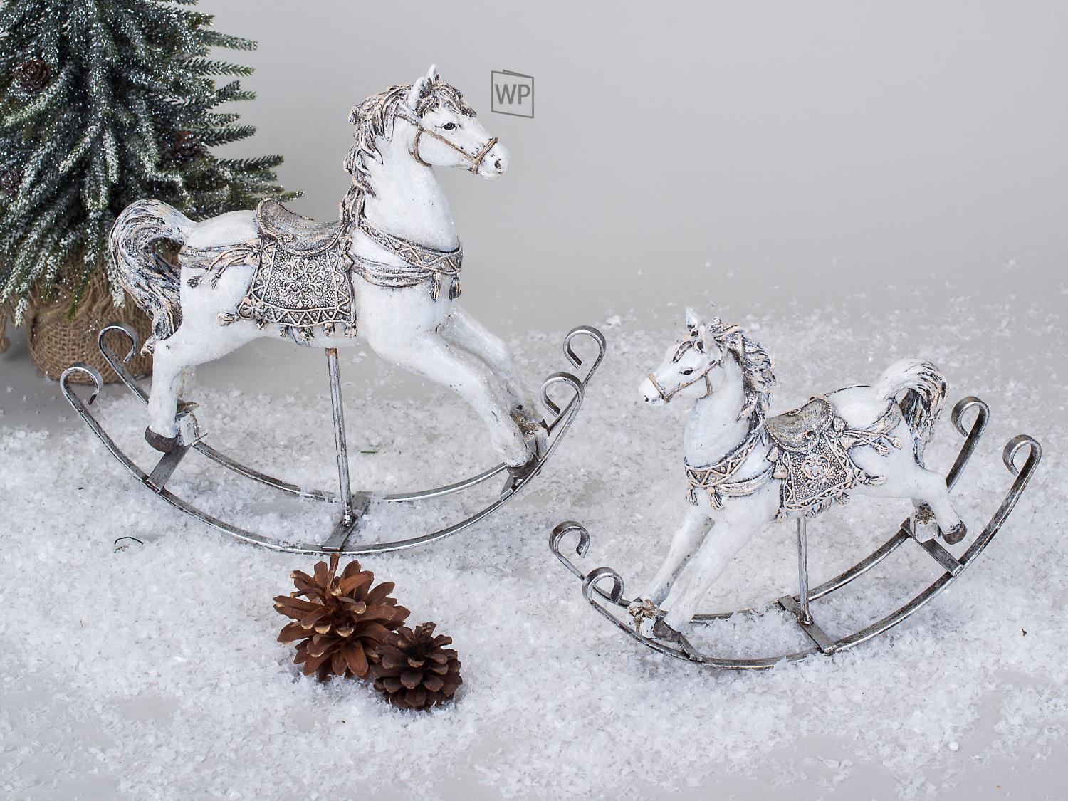 Zwei dekorative Schaukelpferde aus Metall und Holz stehen auf einer schneebedeckten Fläche, umgeben von Tannenzweigen und einer Tannenzapfen.
