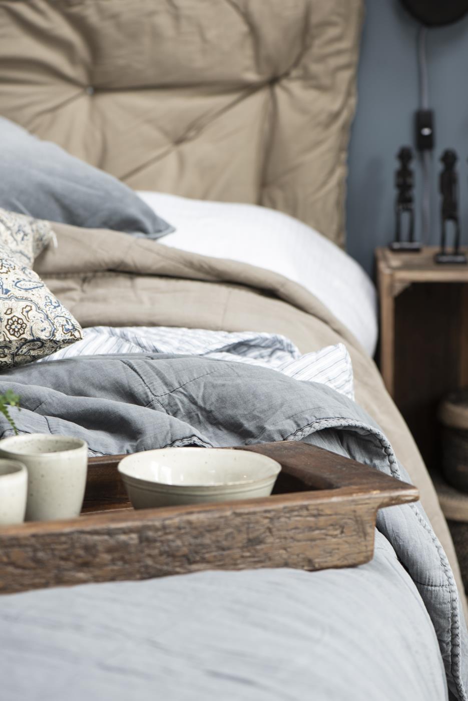 Ein Bett mit beigen und grauen Decken, Kissen und einem Holztablett mit Schalen. Im Hintergrund eine