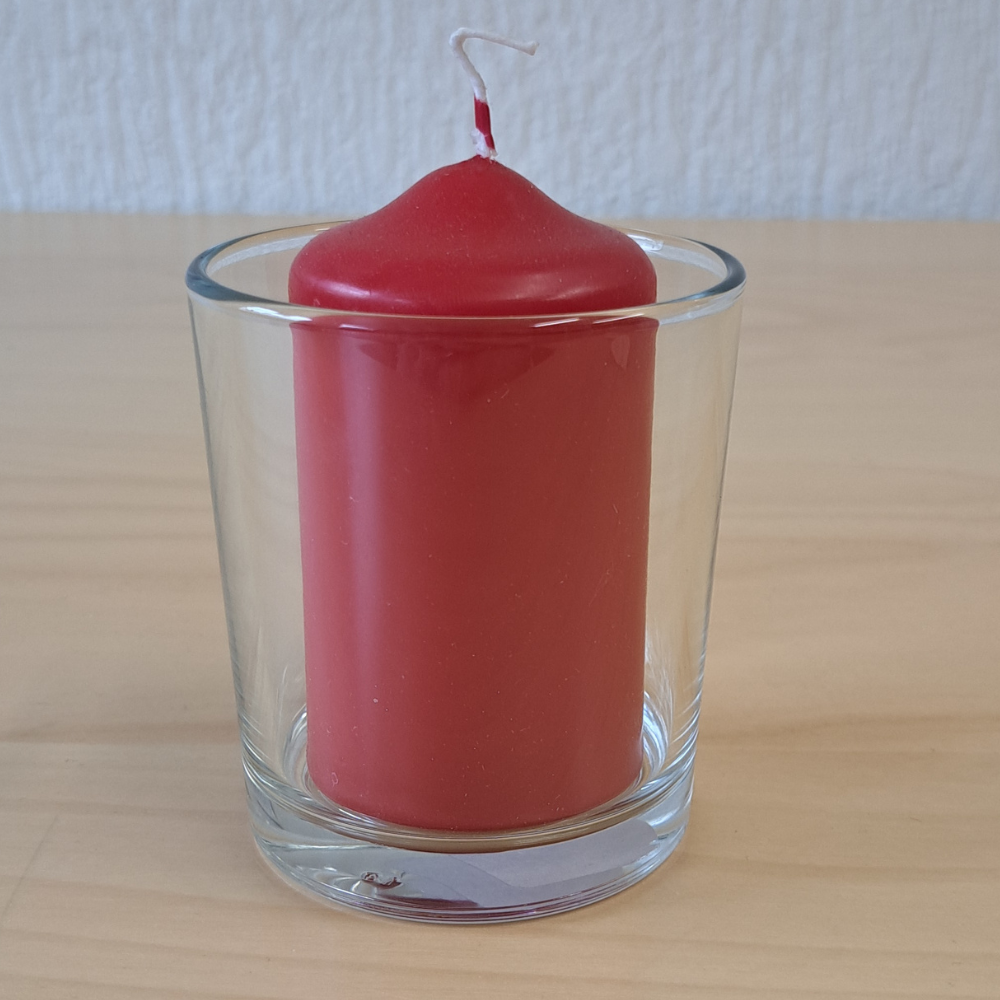 Rote Stumpenkerze in einem klaren Glasbehälter auf einem hellen Tisch.