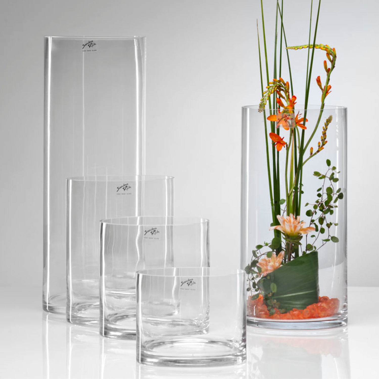 Fünf zylindrische Glasvasen, rechts eine mit orangen Blumen und grünen Stängeln gefüllt