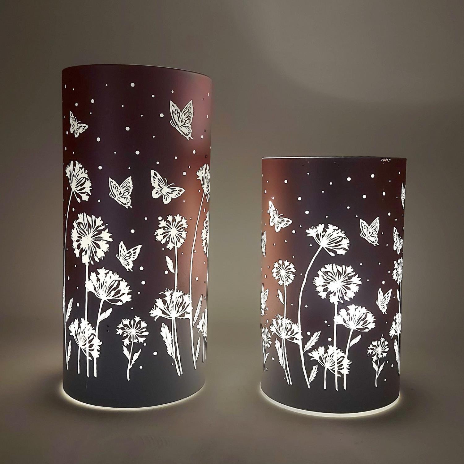 Zwei zylindrische Lampen mit floralem und schmetterlingsartigem Muster auf dunklem Hintergrund, beleuchtet von innen.