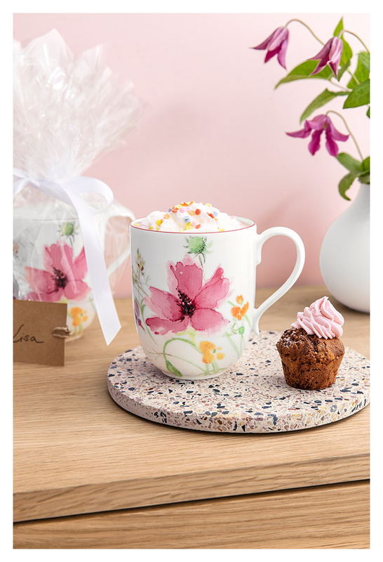 Eine Tasse mit Blumenmuster steht auf einem Untersetzer. Daneben ein kleiner Muffin mit rosa Topping