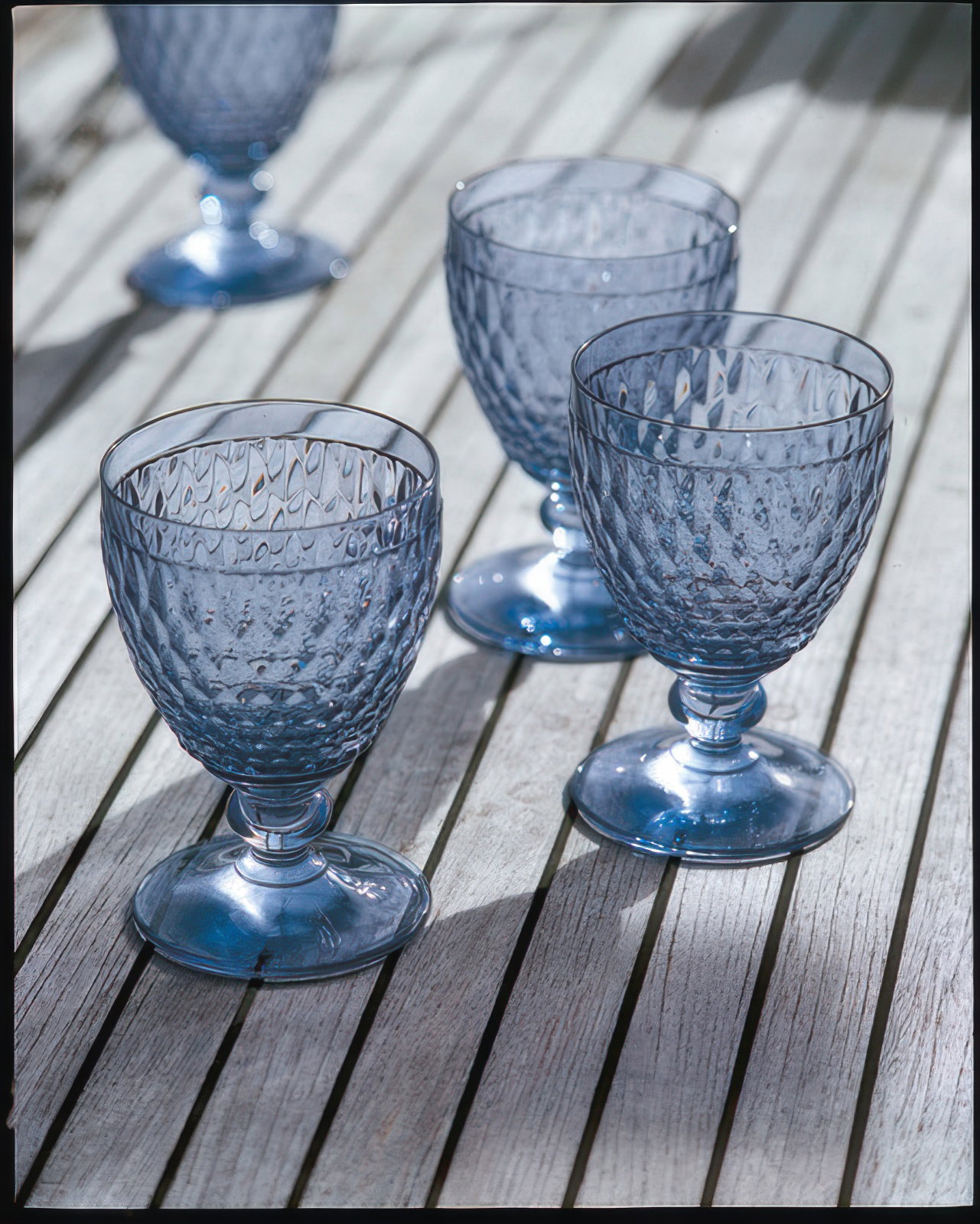 Vier blaue Gläser mit strukturiertem Muster stehen auf einem Holztablett. Die Gläser haben einen Fuß und sind leicht transparent.