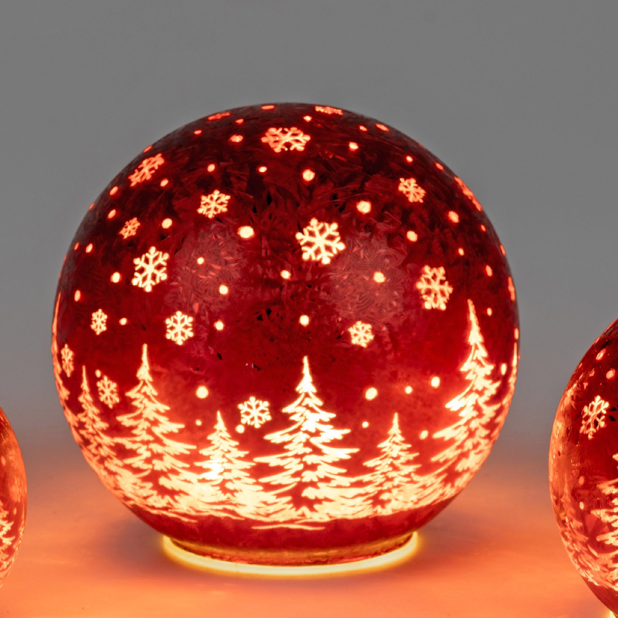 Runde, rote Weihnachtskugel mit weißen Schneeflocken und Tannenbäumen, die von innen beleuchtet ist.