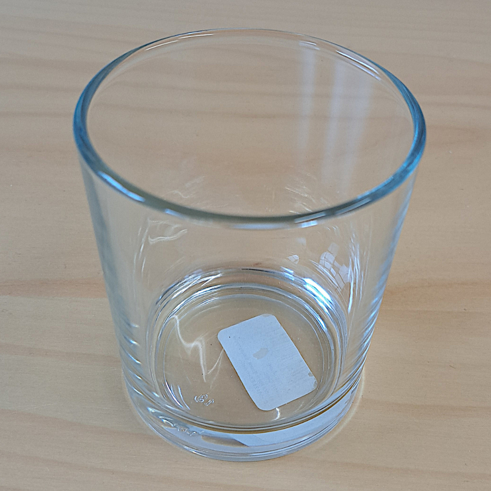 Ein transparenter Glasbecher steht auf einem hellen Tisch. Im Inneren liegt ein kleines, weißes Etik