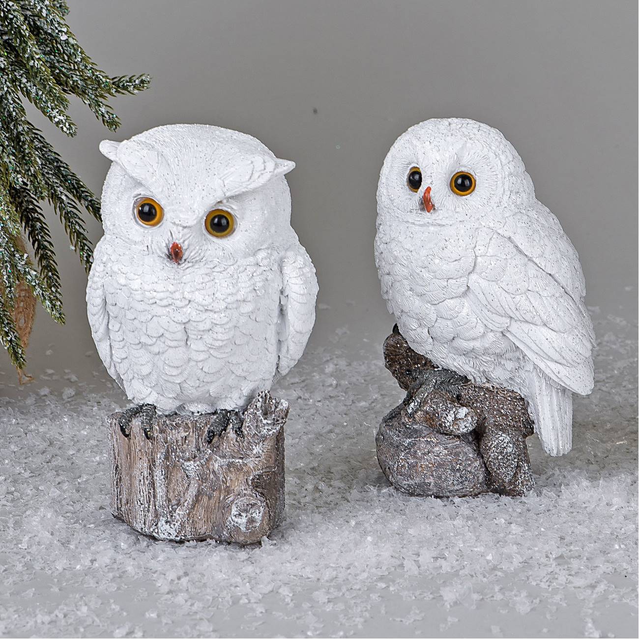 Zwei weiße Eulenfiguren stehen auf Holzstücken, umgeben von Kunstschnee und einer grünen Pflanze im