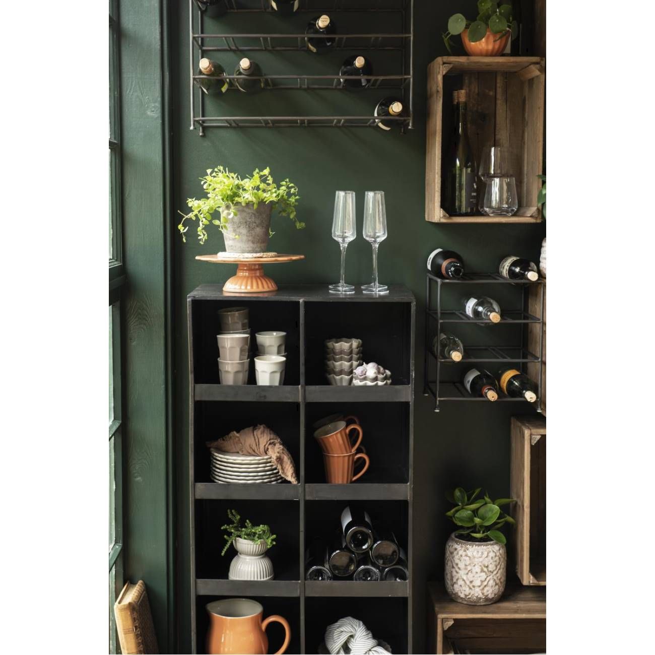 Regal mit Geschirr, Gläsern und Pflanzen. An der Wand hängen Weinflaschen. Dunkle Wandfarbe und Holzregale sind sichtbar.