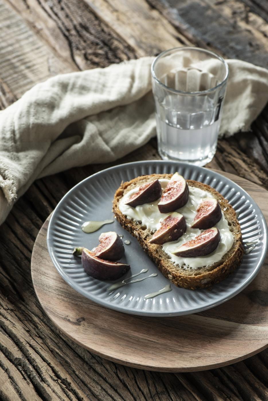 Ein Teller mit einem Stück Brot, belegt mit Frischkäse und Feigen, daneben ein Glas Wasser und ein Tuch auf einem Holztisch.