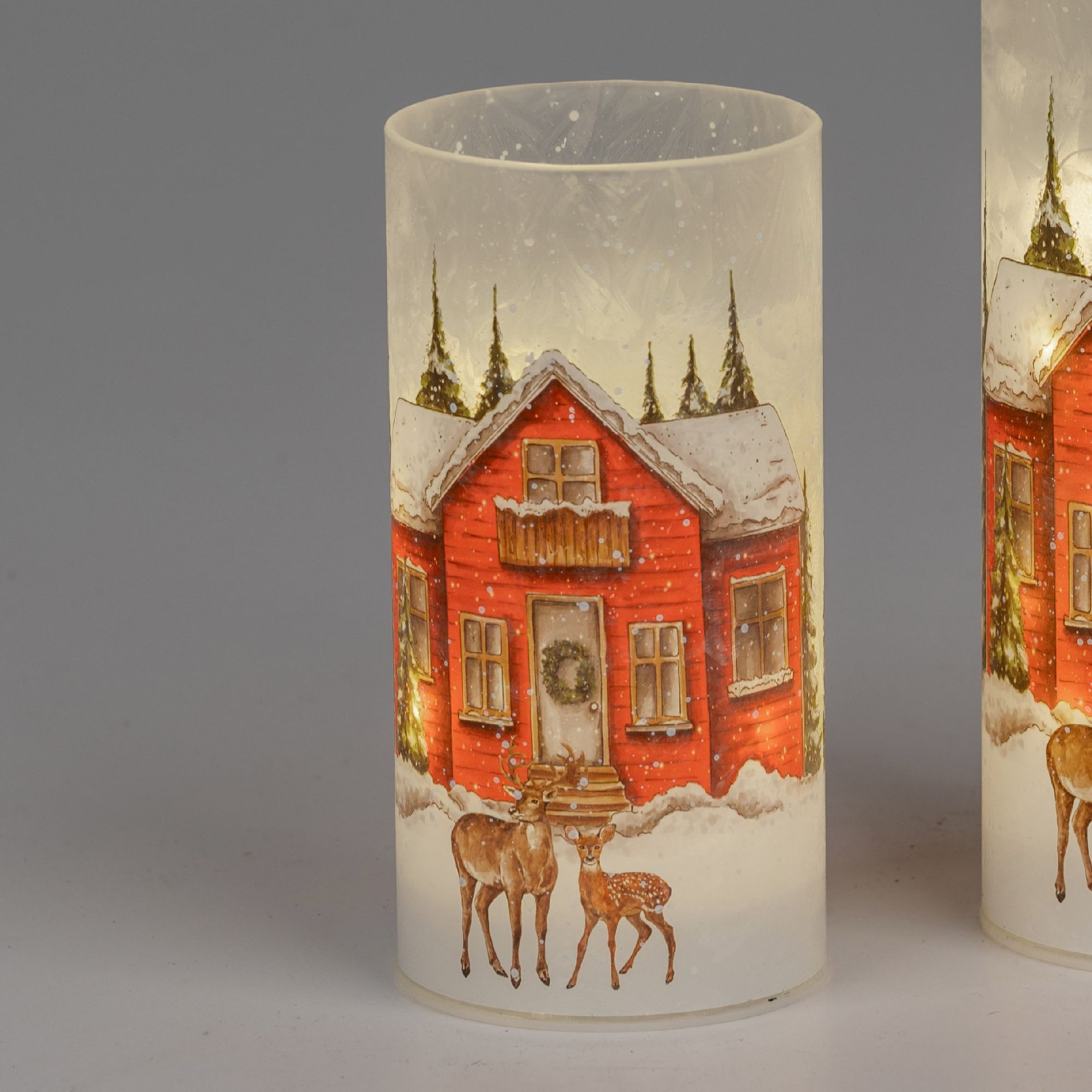 Zwei leuchtende Kerzen mit winterlicher Motive: ein rotes Haus, Tannenbäume und zwei Rehe im Schnee.